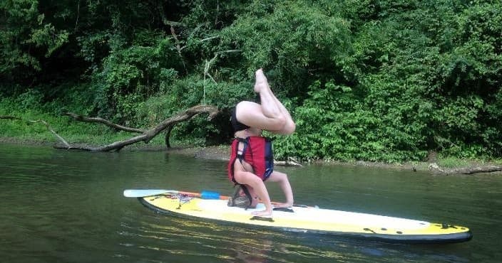 Person doing SUP yoga headstand on paddleboard near green shore