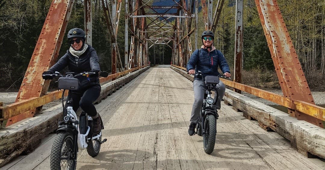 Two riders on e-bikes crossing a rustic bridge through forest