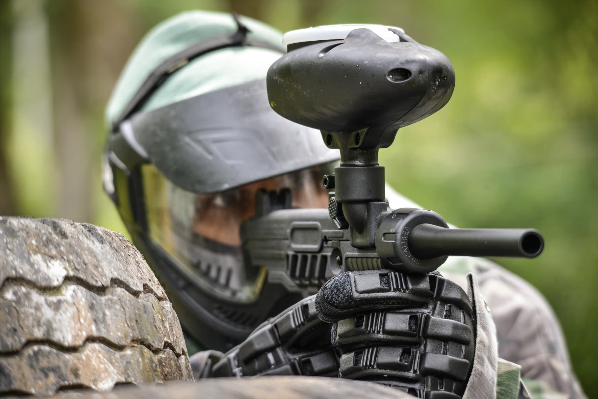 Paintball player aiming from behind tire in woodland field