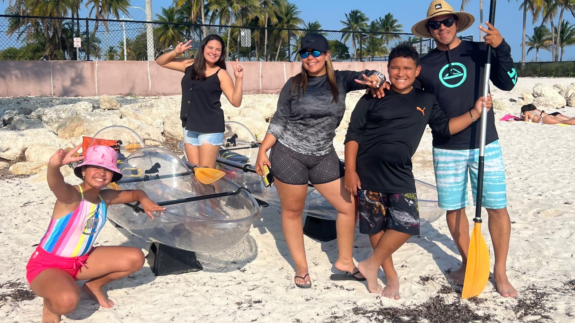 Group posing with transparent kayaks on sunny beach near palms