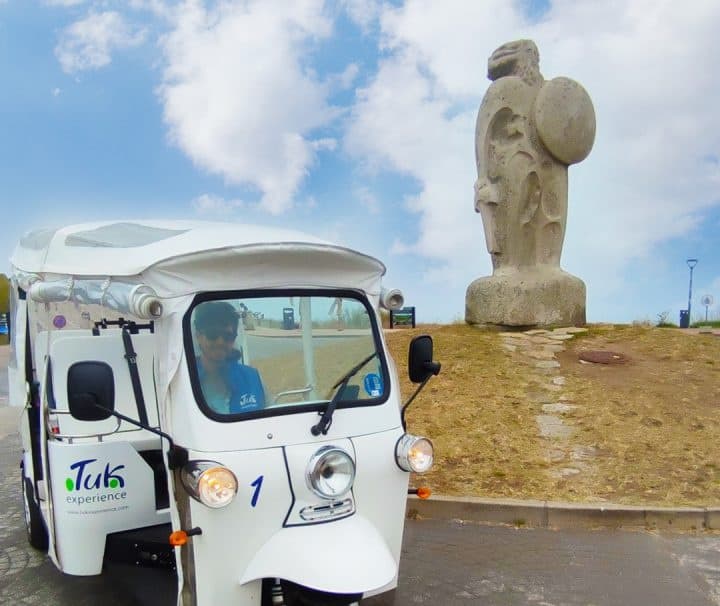 White tuk-tuk parked by coastal statue with driver visible on cloudy day