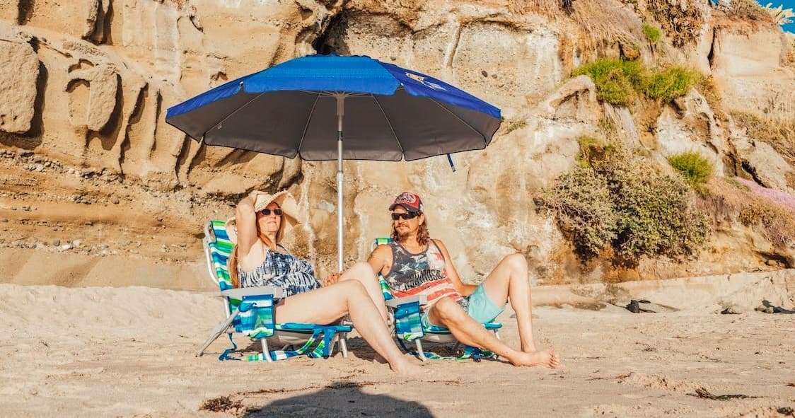 Two people relaxing under beach umbrella by coastal cliffs and sand