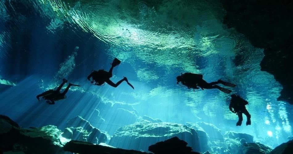 Silhouetted scuba divers in sunlit underwater cave with rays