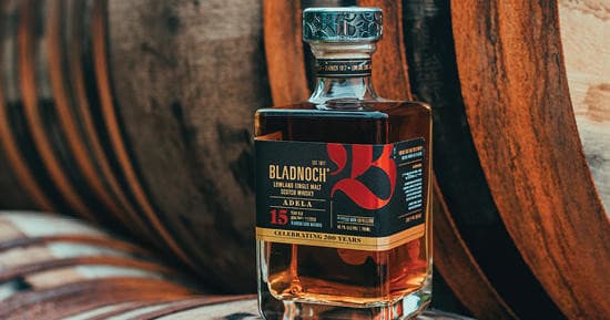 Bladnoch whisky bottle by stacked barrels outdoors in warm sunlight