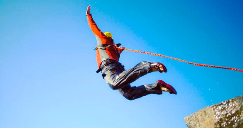 Person cliff-jumping with safety rope over rocky ledge and blue sky