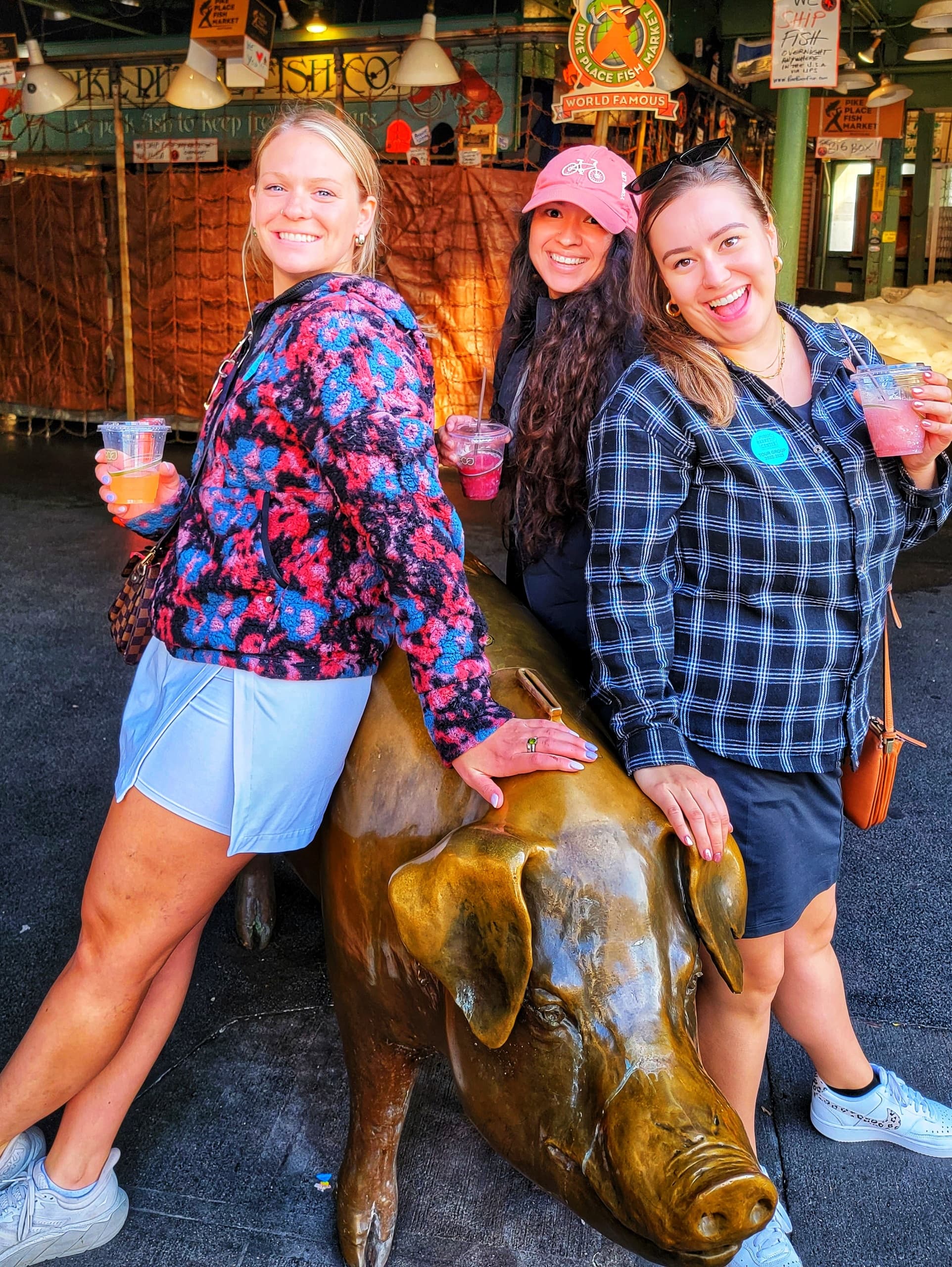 Women posing on bronze pig statue at urban market with drinks