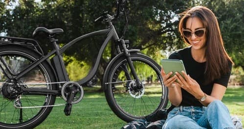 Woman with e-bike relaxing on green park lawn, checking tablet