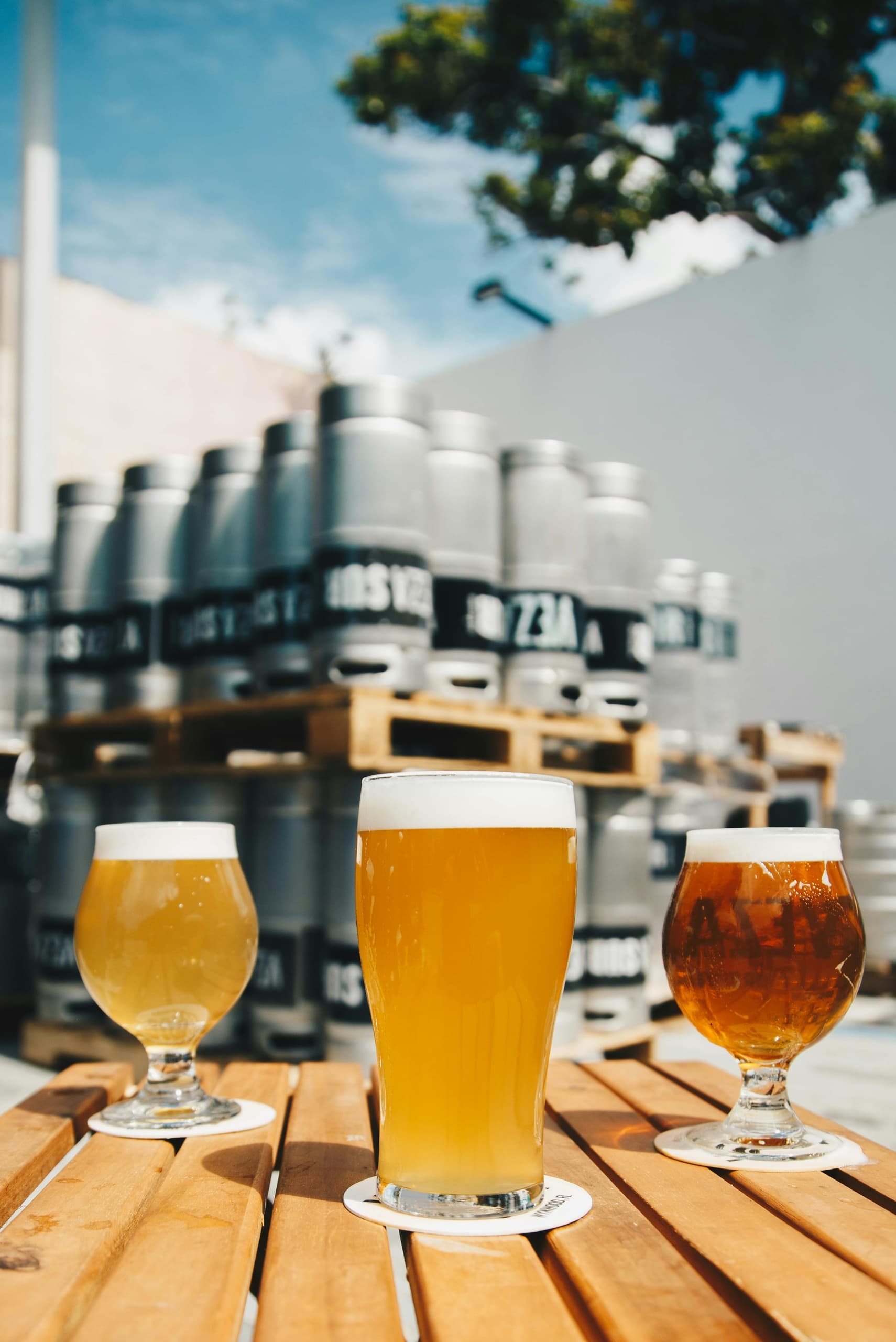 Three craft beer glasses on outdoor table with brewery kegs