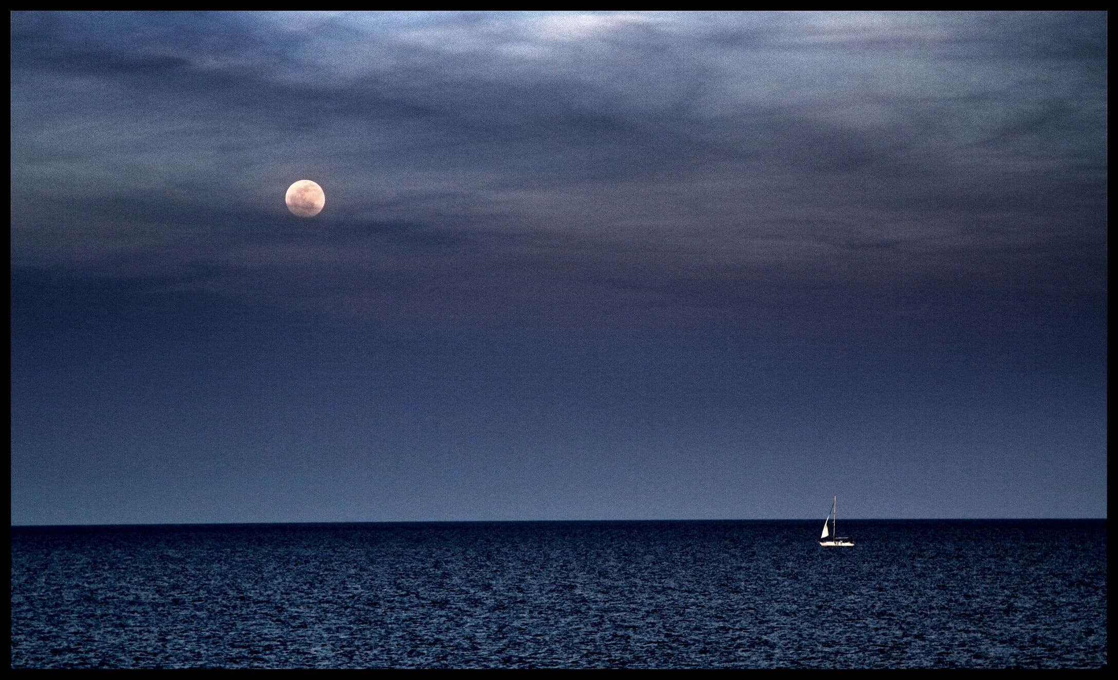 Small sailboat under moonlit sky on calm open ocean at dusk