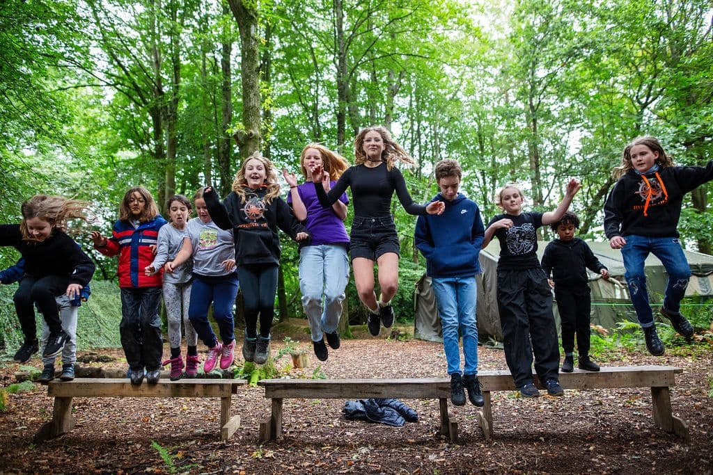 Young campers jumping across log obstacle in leafy woodland