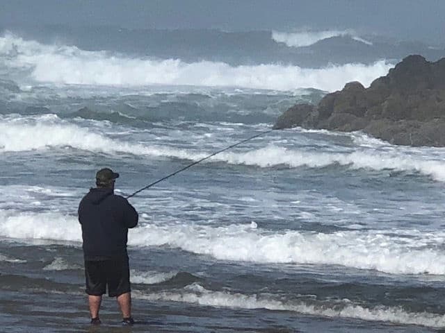 Angler surfcasting on rocky Pacific shore with dramatic waves