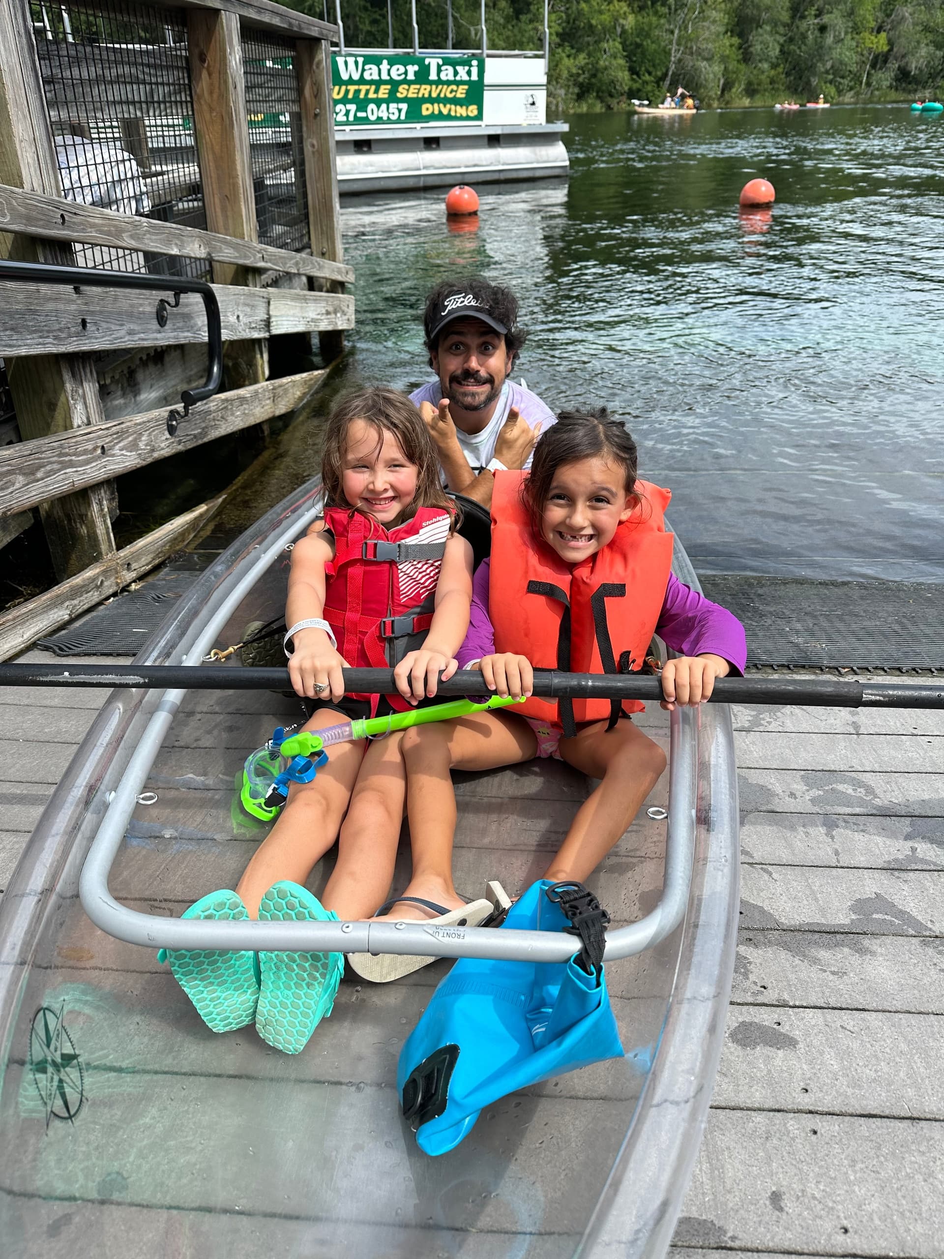 Family paddling a clear kayak at a spring-fed river dock, smiling