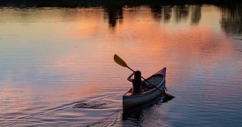 Solo kayaker paddling at sunset on calm river, reflective waters