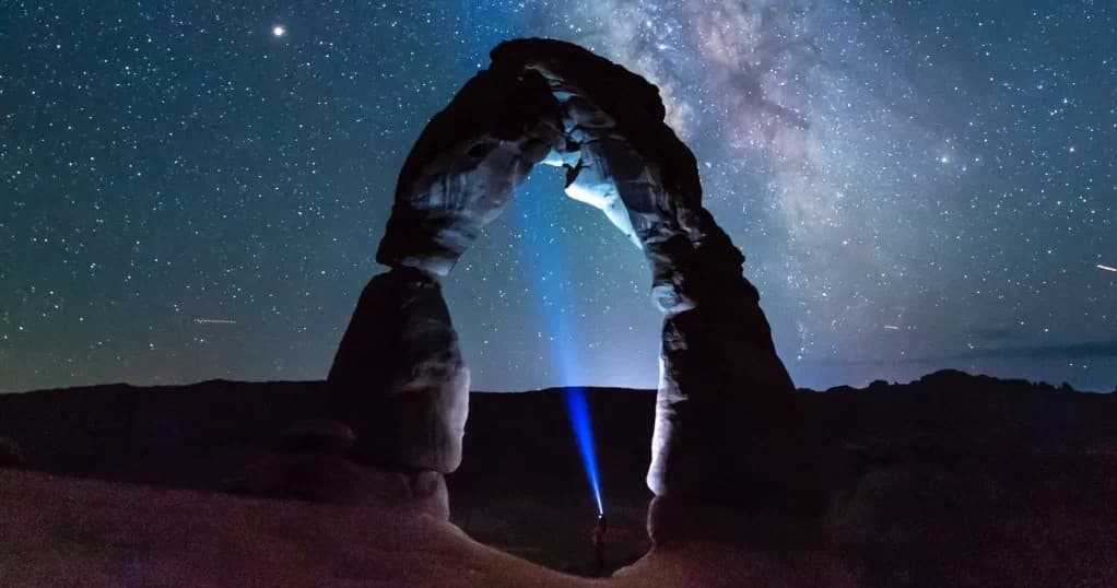 Hiker shining blue beam under Delicate Arch beneath Milky Way