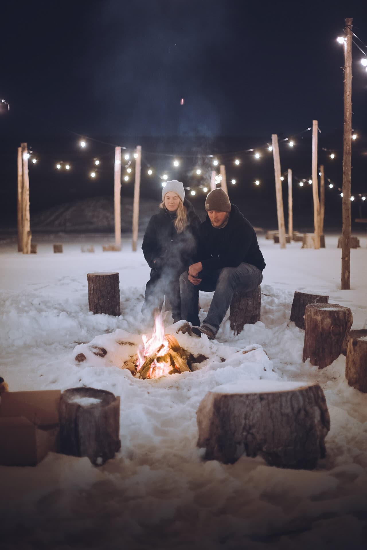 Couple warming by campfire at snowy outdoor farm with string lights
