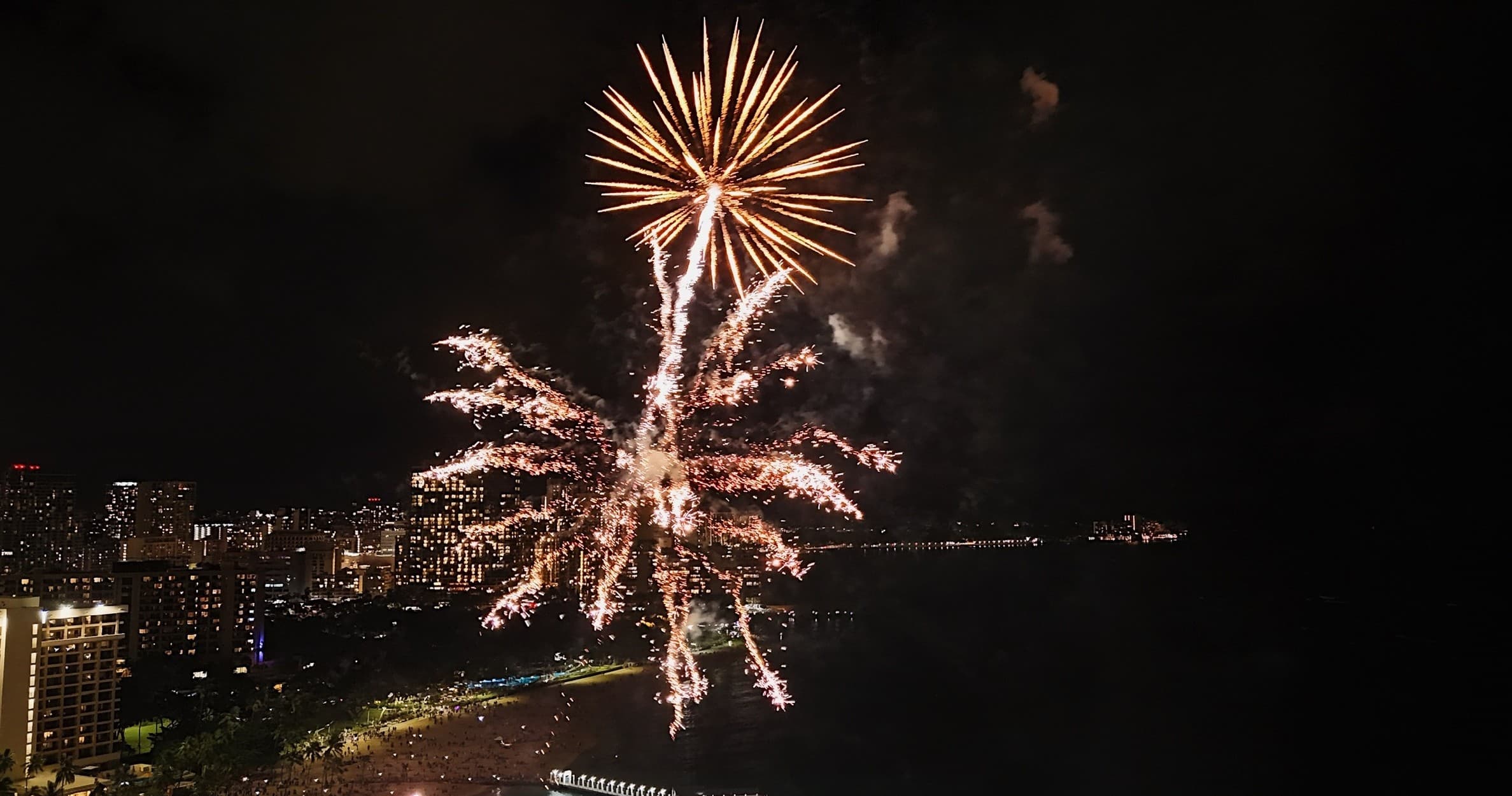 Nighttime fireworks over Waikiki beach shoreline, city lights
