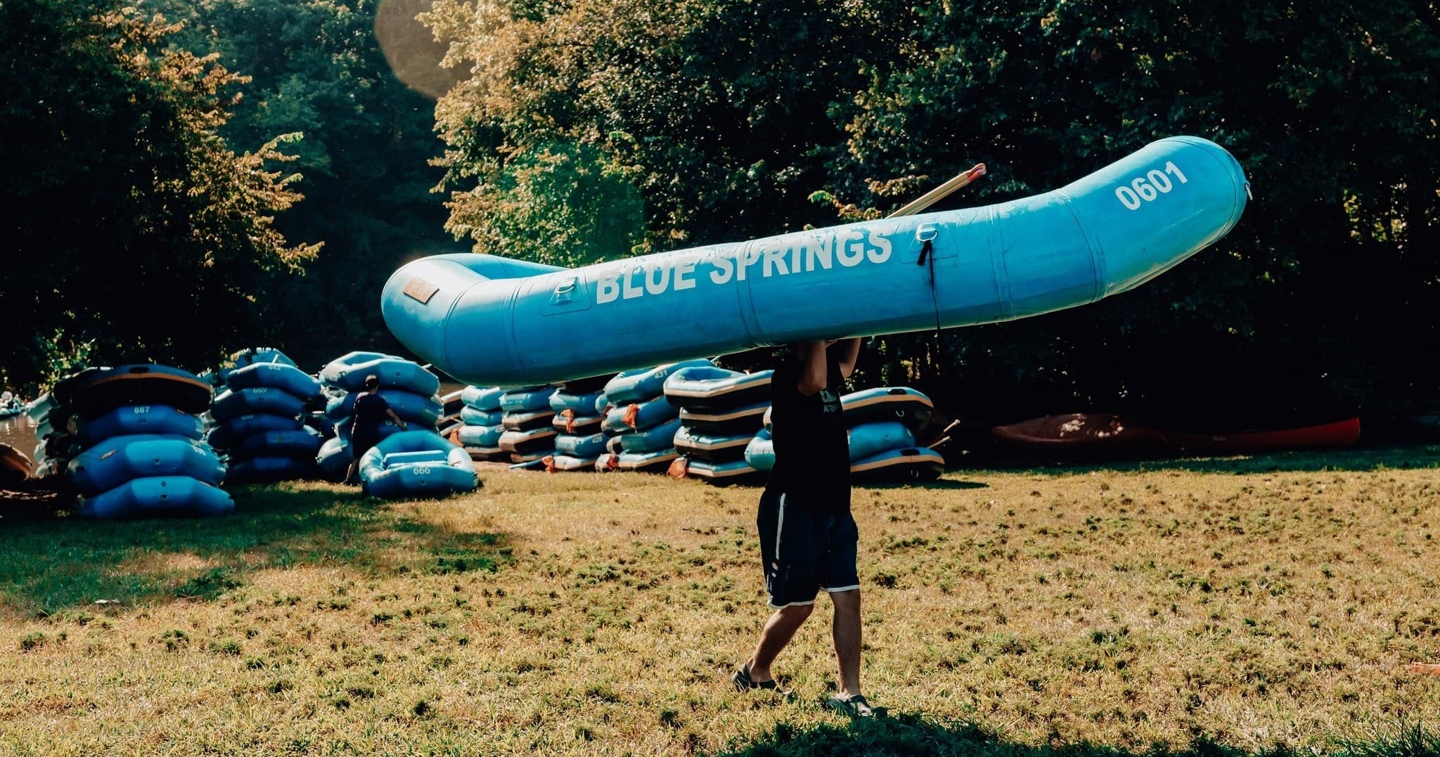 Staff carrying inflatable raft across grassy staging area at river outfitter