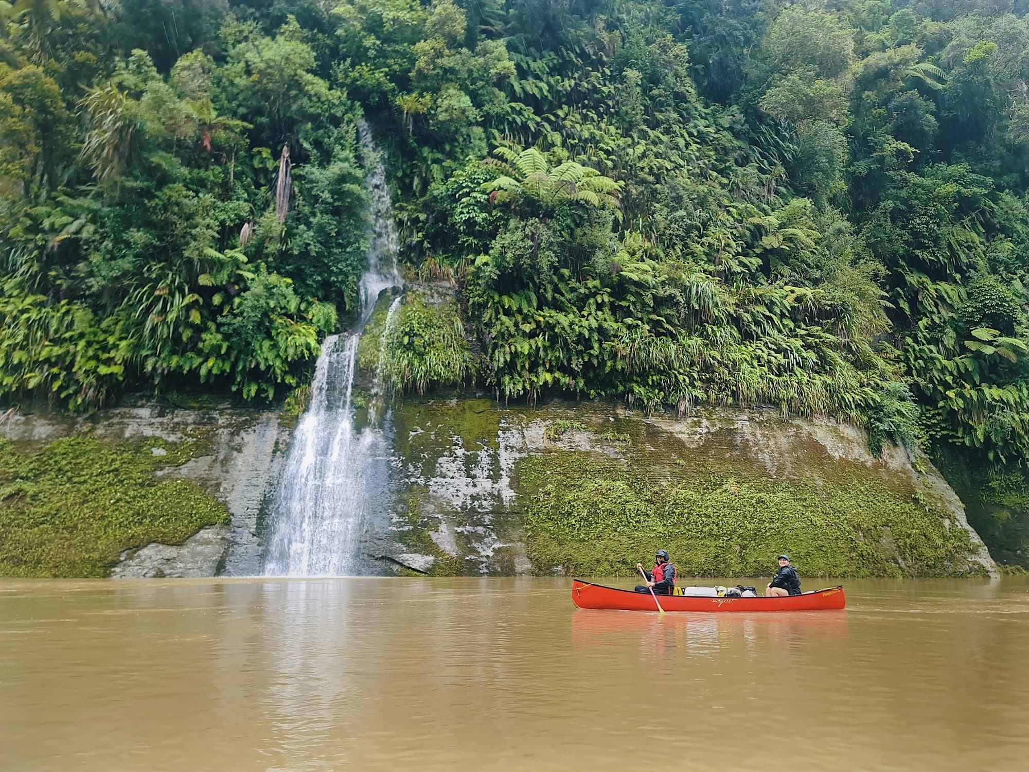 Red canoe passing waterfall on tropical river with lush cliffs