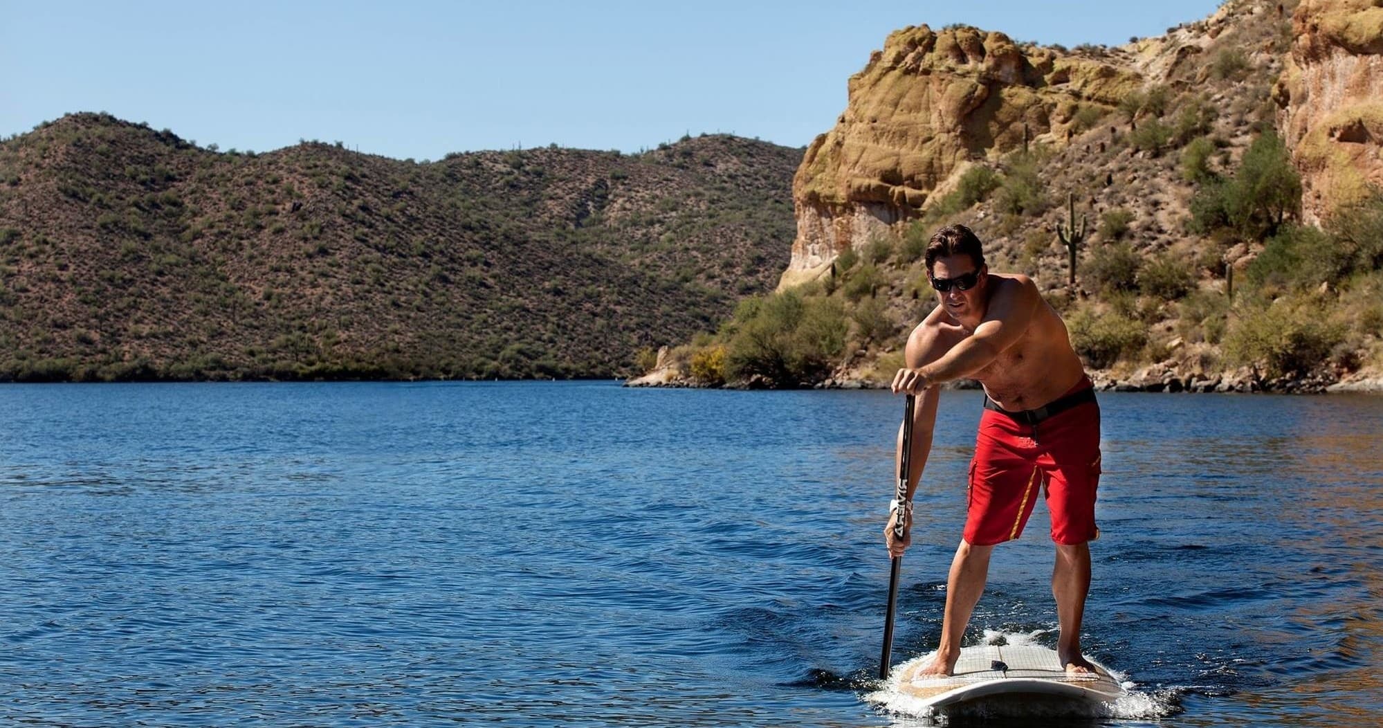 Paddleboarder near rocky shoreline with desert hills backdrop