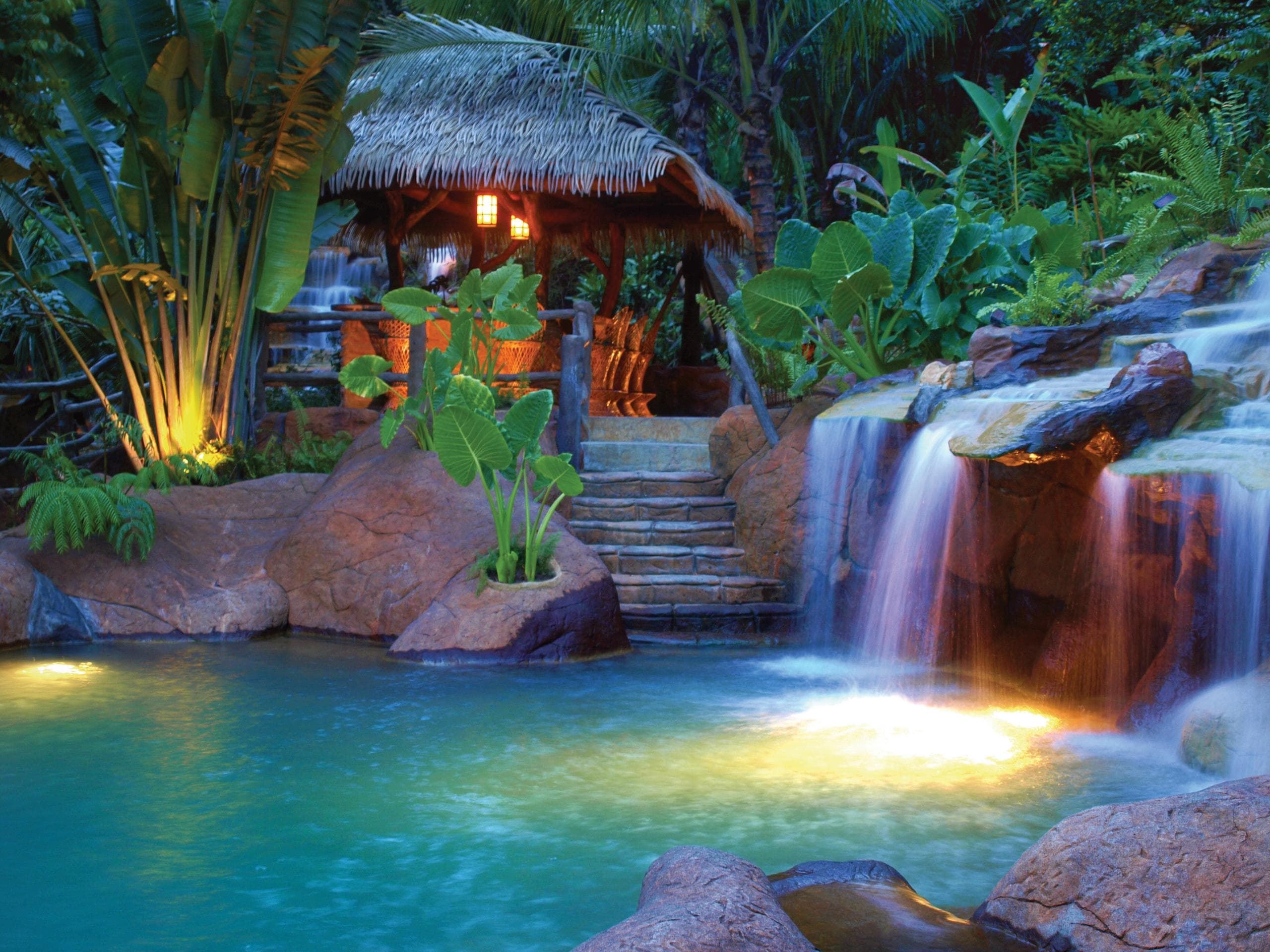 Tropical resort pool with lit waterfall and lush garden at dusk