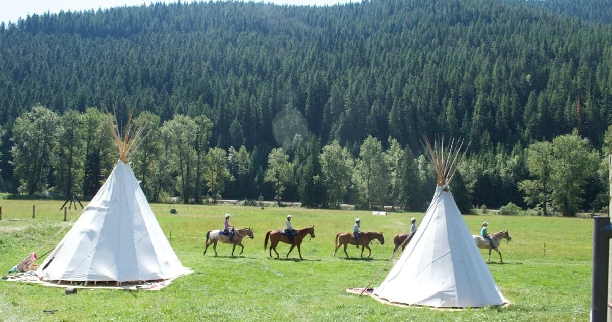 Horseback riders passing white teepees on grassy ranch by forest