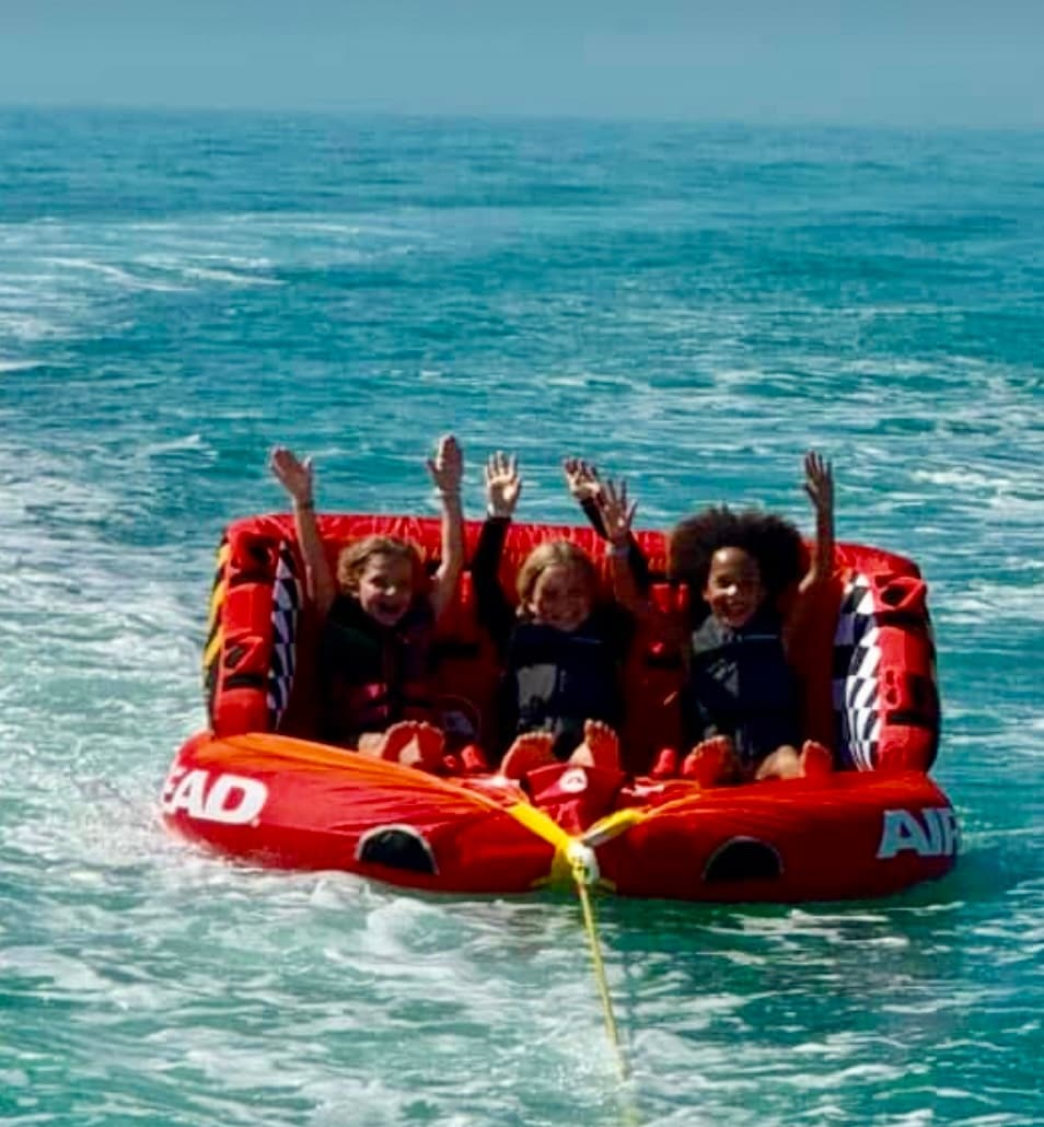 Three riders tubing behind boat on choppy turquoise ocean, hands raised