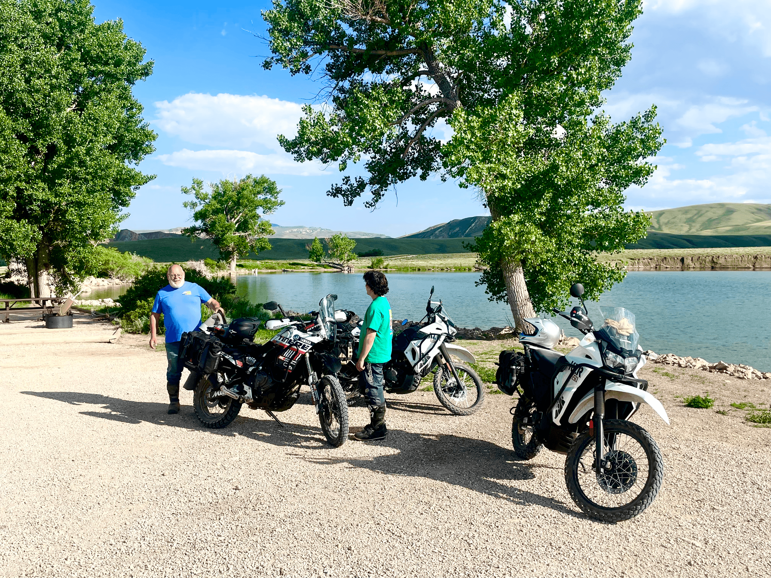 Adventure motorcycles parked lakeside with riders beside prairie reservoir