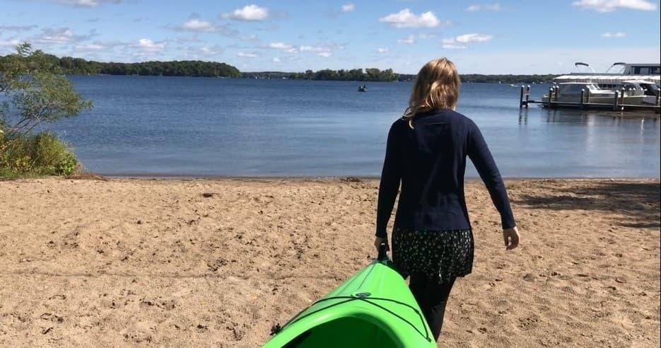 Person carrying green kayak toward sandy lake beach on calm day