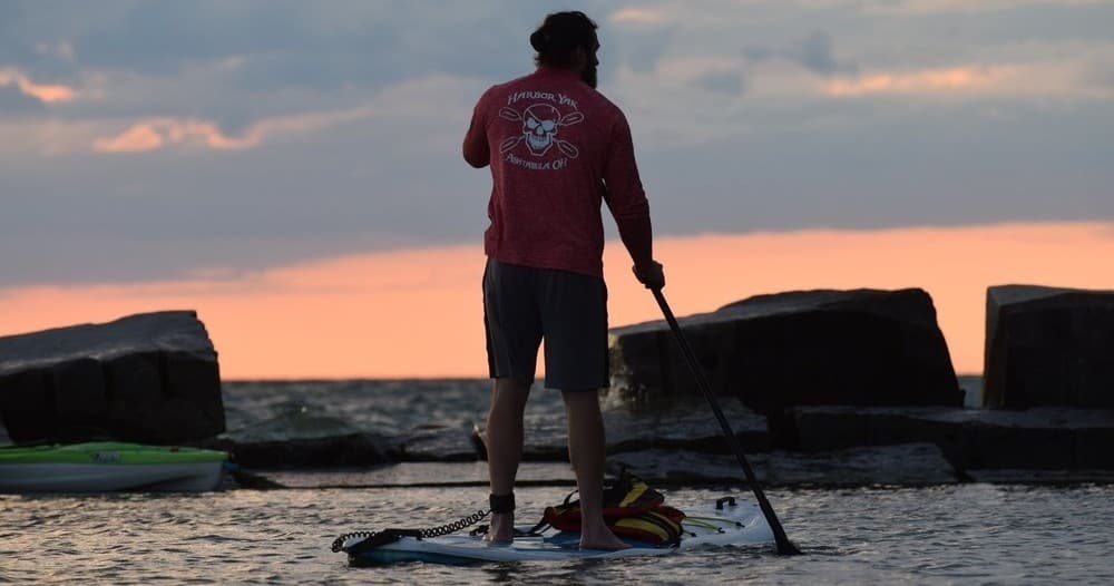 Stand-up paddleboarder silhouetted by orange sunset near coastal rocks