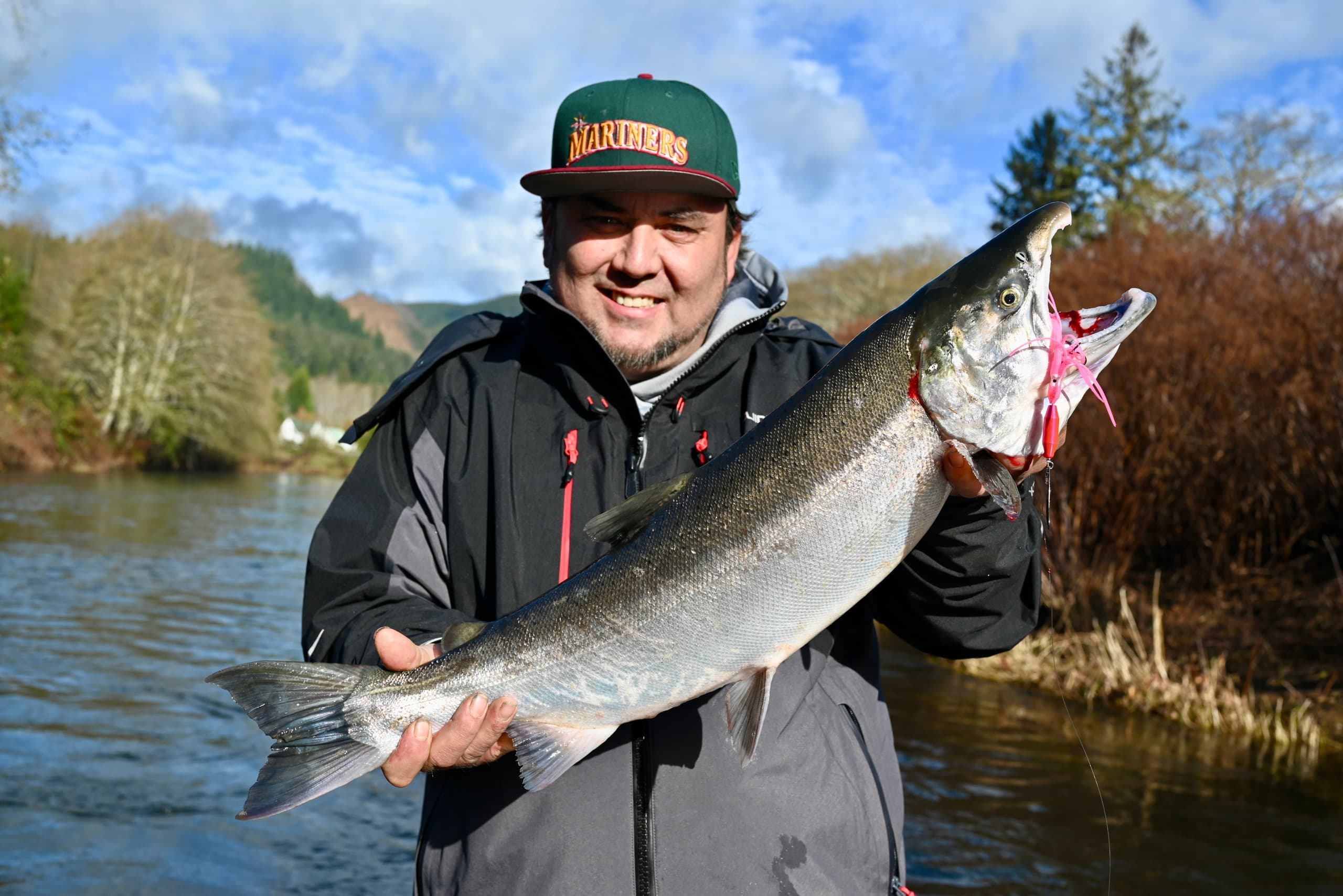 Smiling angler holding steelhead by river under blue sky