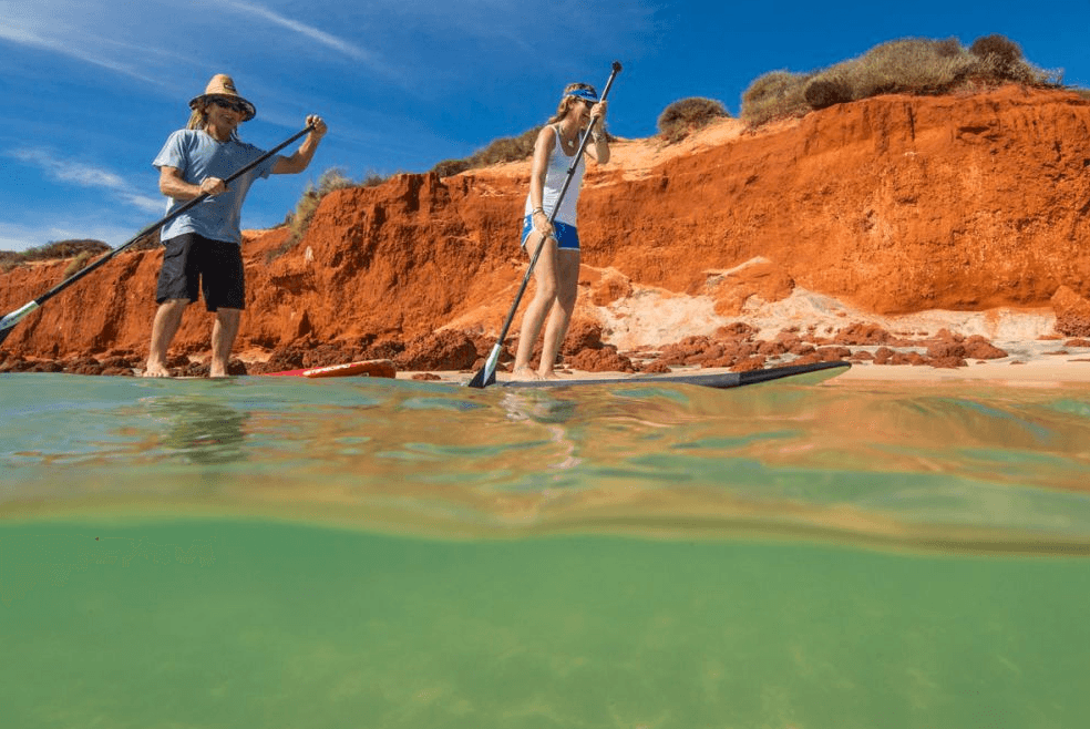 Stand-up paddleboarding by red cliffs in shallow green water