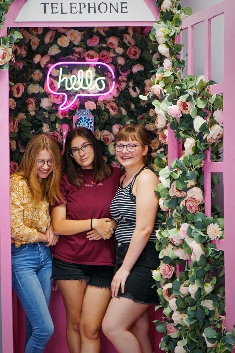 Three friends posing inside pink floral selfie booth with neon