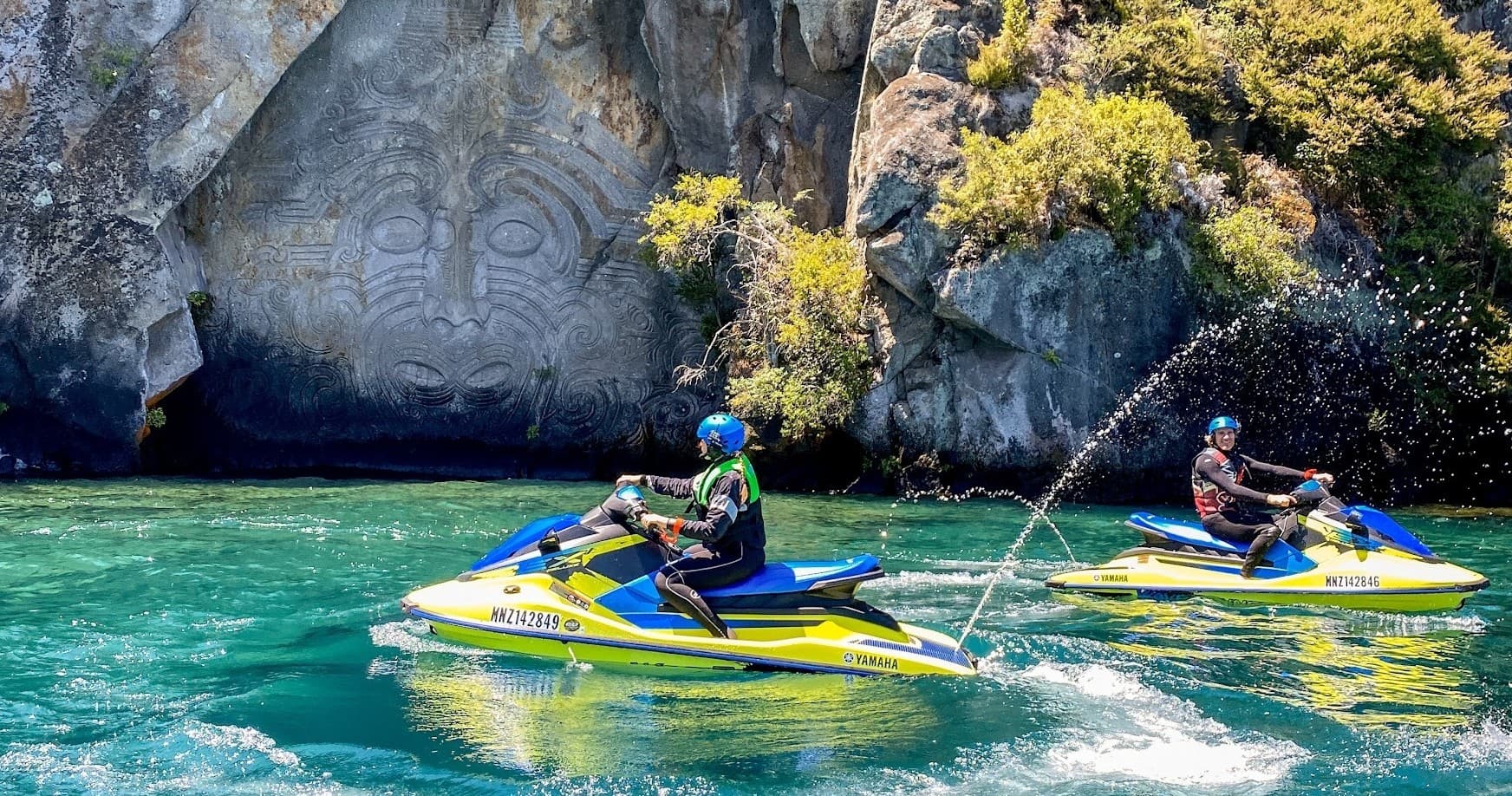 Jet ski riders racing on turquoise lake past rocky cliff