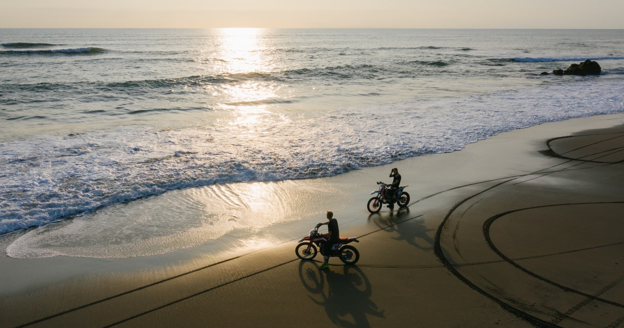 Two riders on beach motorcycles at sunset carving tracks on sand