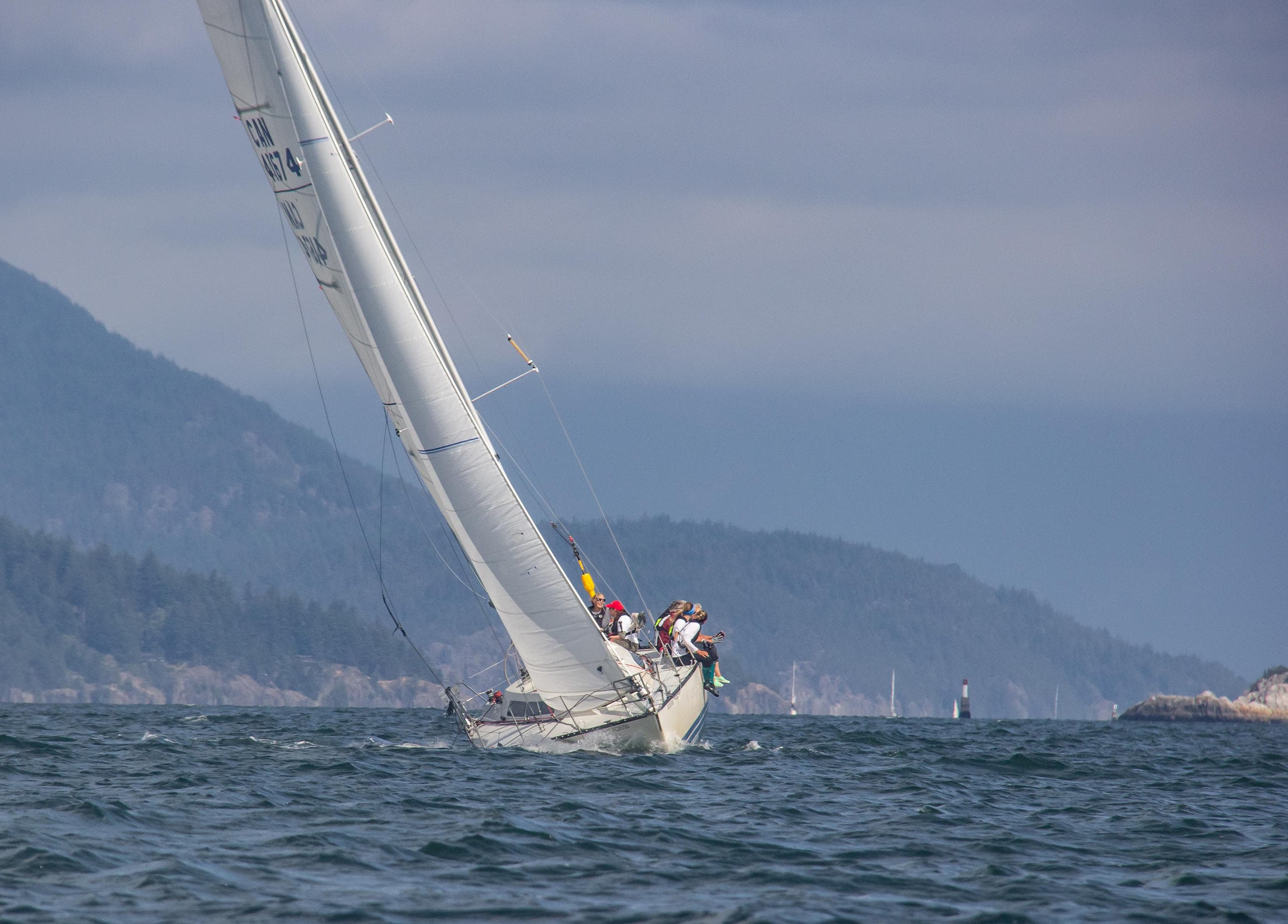 Racing sailboat heeling off rugged coastline under cloudy sky