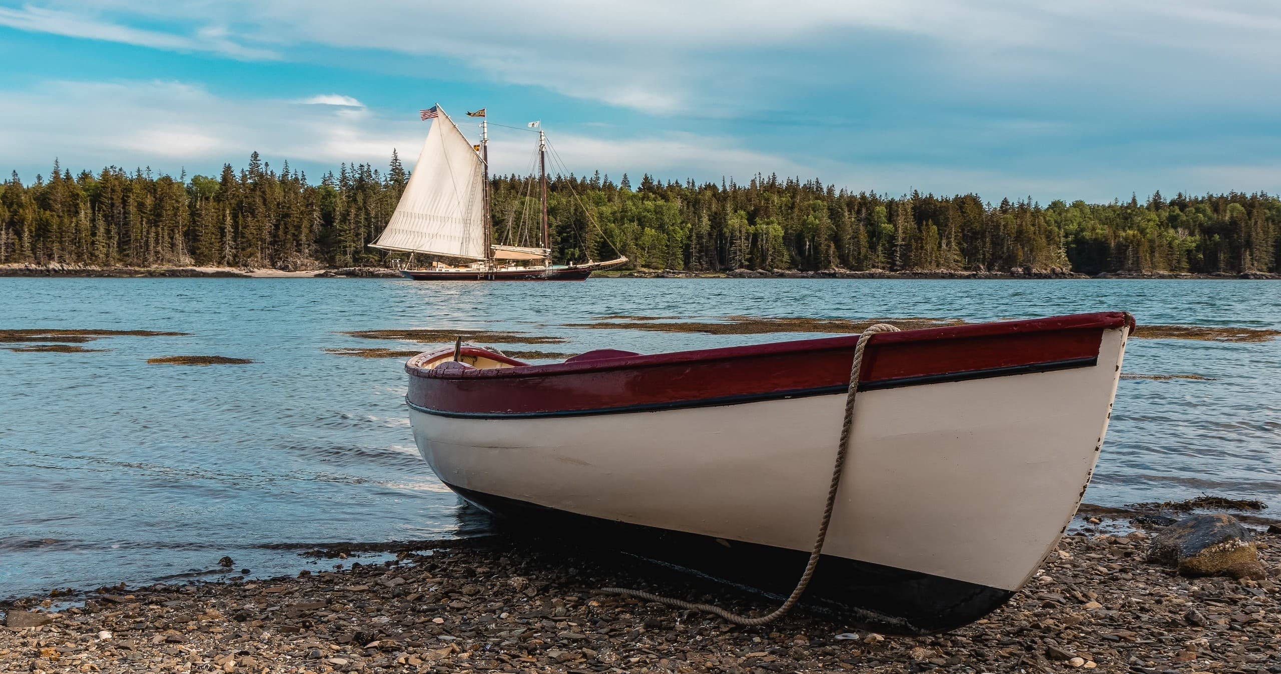 Rowboat on rocky Maine shore with sailboat cruising offshore