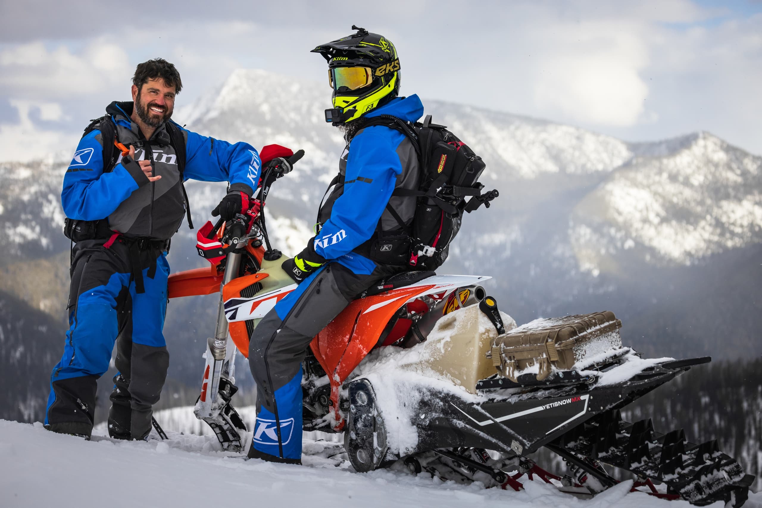 Two riders prepping snowbike with mountain vista backdrop