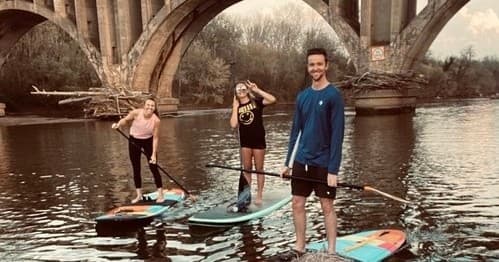 Three friends stand on paddleboards near arched bridge at dusk