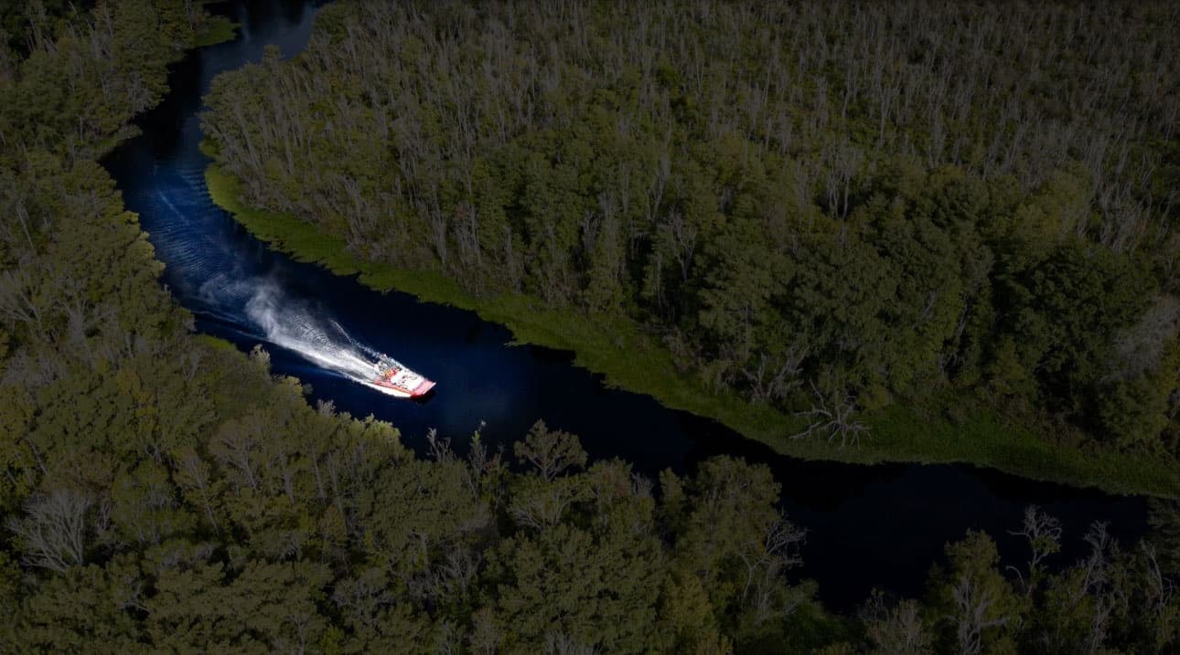 Airboat slicing narrow dark river through dense Florida wetland forest
