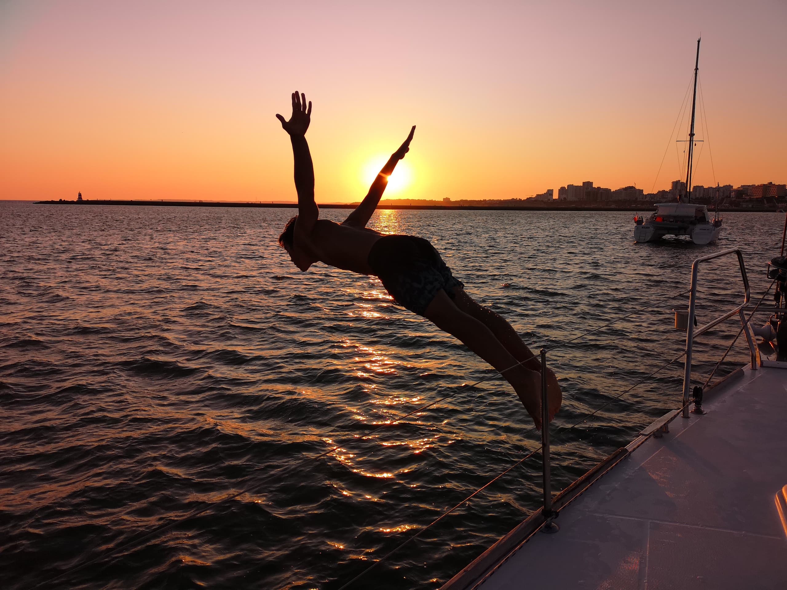 Person diving off sailboat into sunset sea near coastline