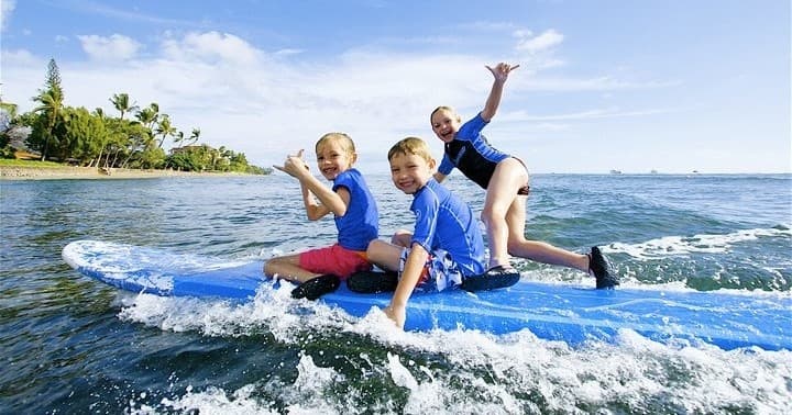 Three kids balancing on a blue surfboard in shallow tropical surf