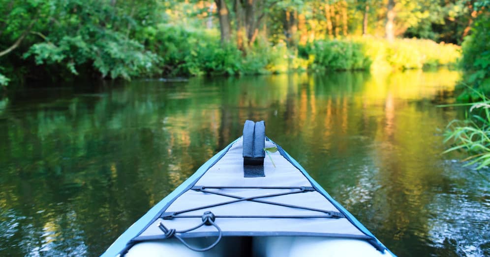 Front of blue kayak navigating calm forest river at sunset
