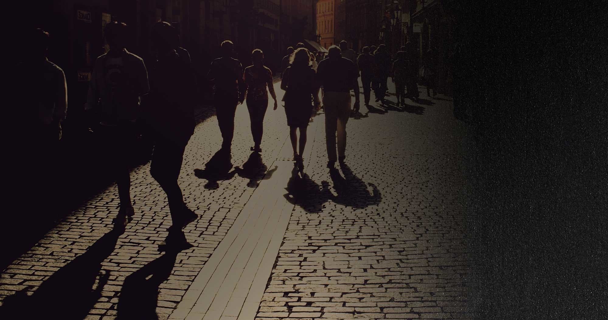 City pedestrians casting long shadows walking on cobblestone street