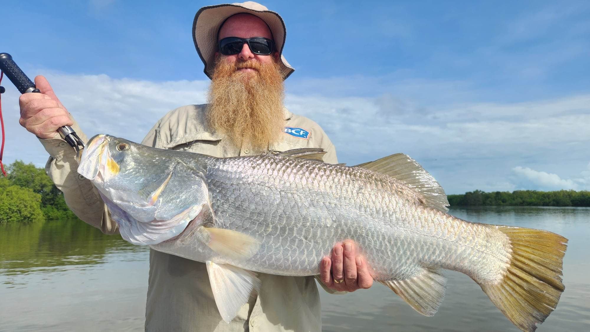 Angler holding trophy barra on calm river estuary, tropical sky