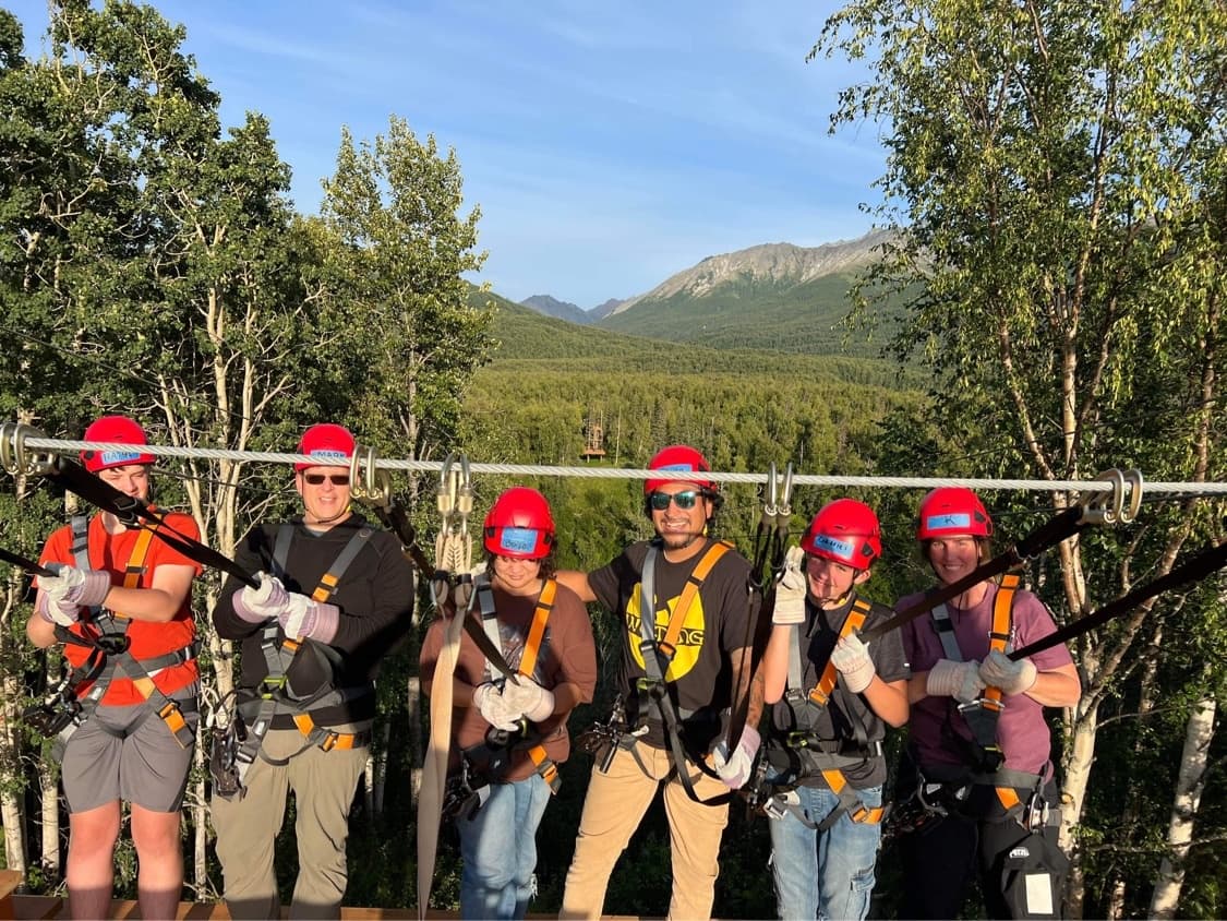 Zipline crew wearing helmets and harnesses above forested valley