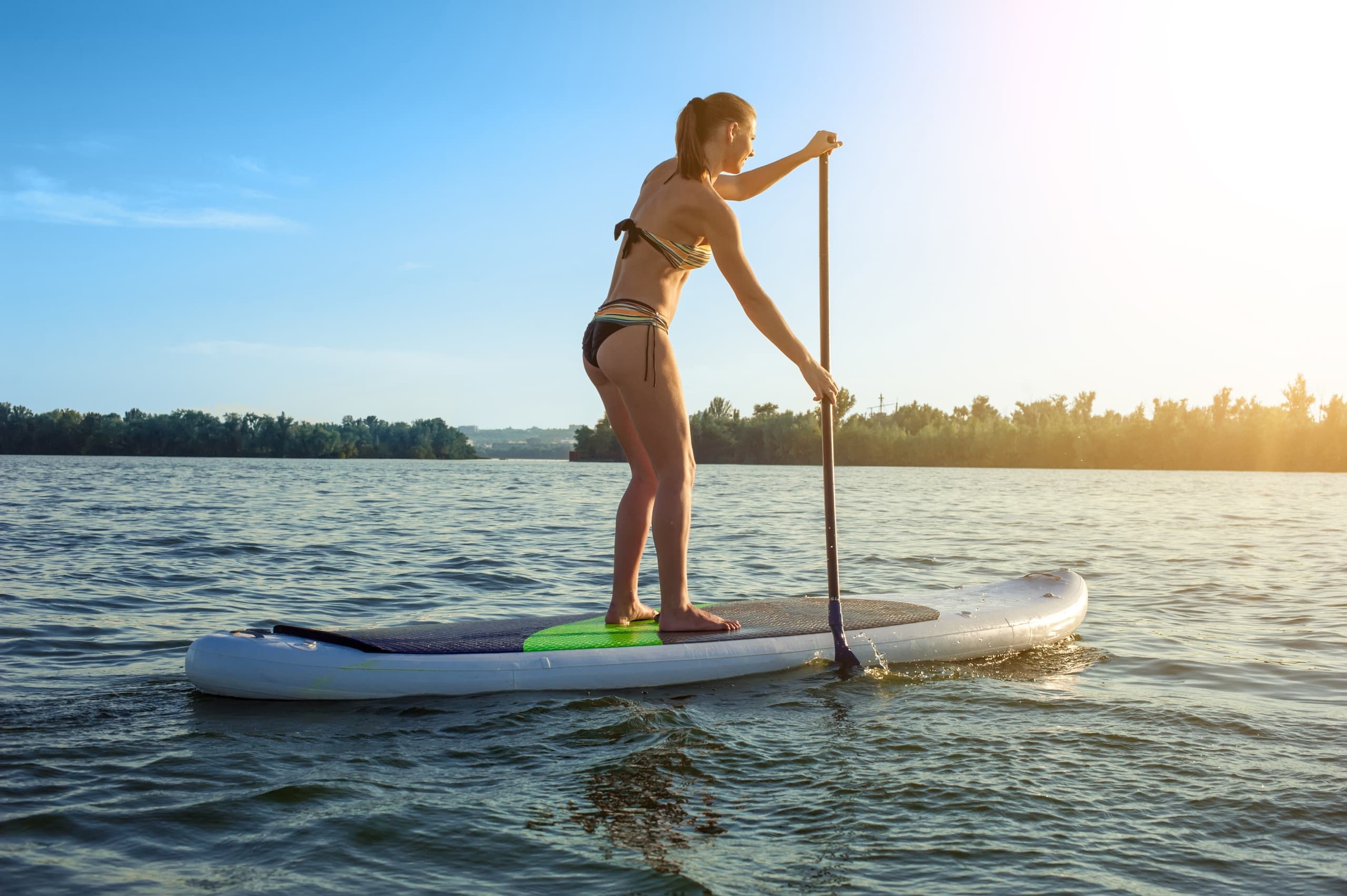 Woman paddleboarding on open lake with bright sky and sun