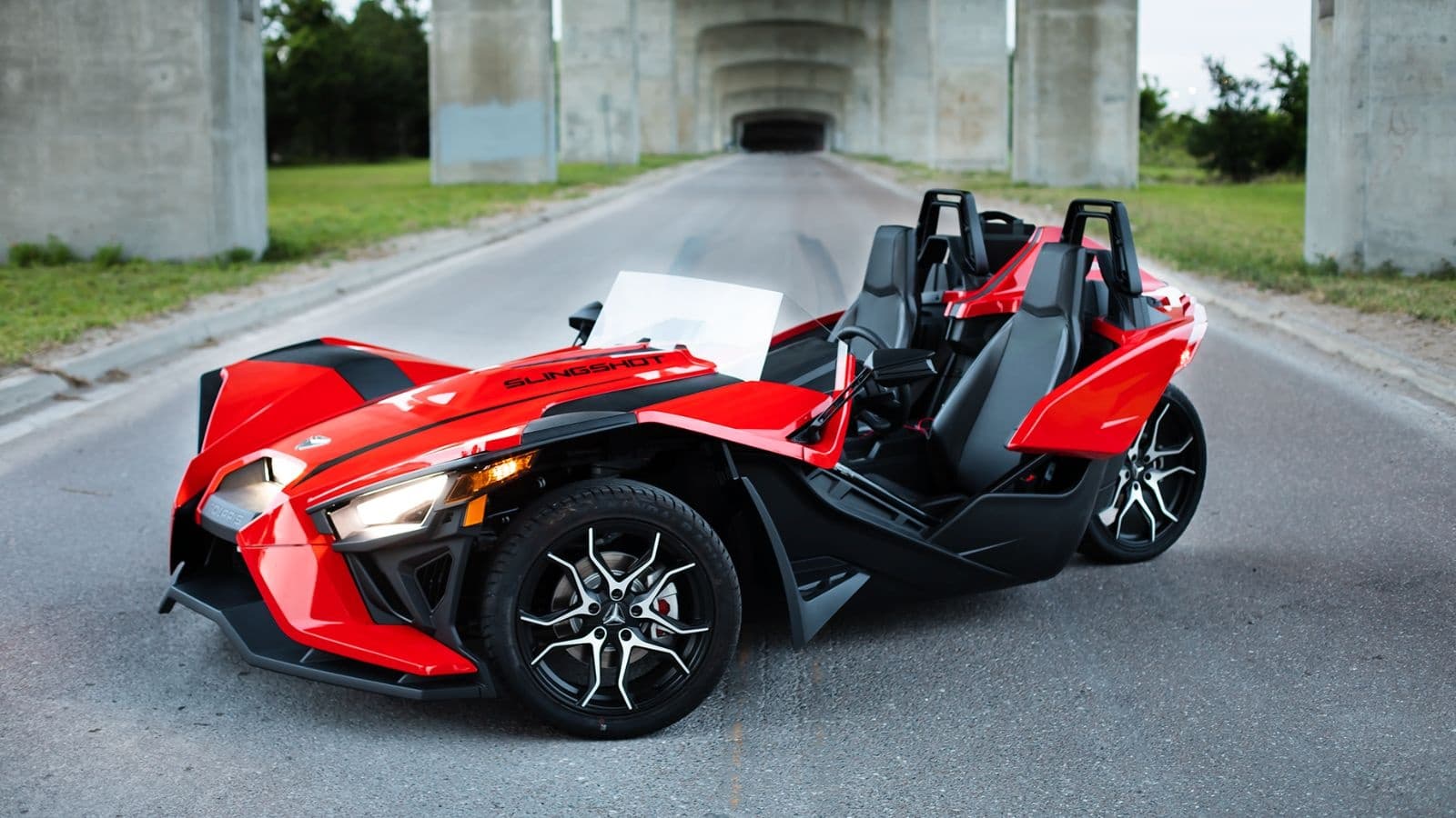 Red open-air Slingshot parked on coastal road near waterfront bridge