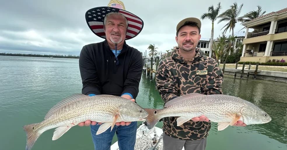 Two anglers holding redfish on boat with waterfront homes