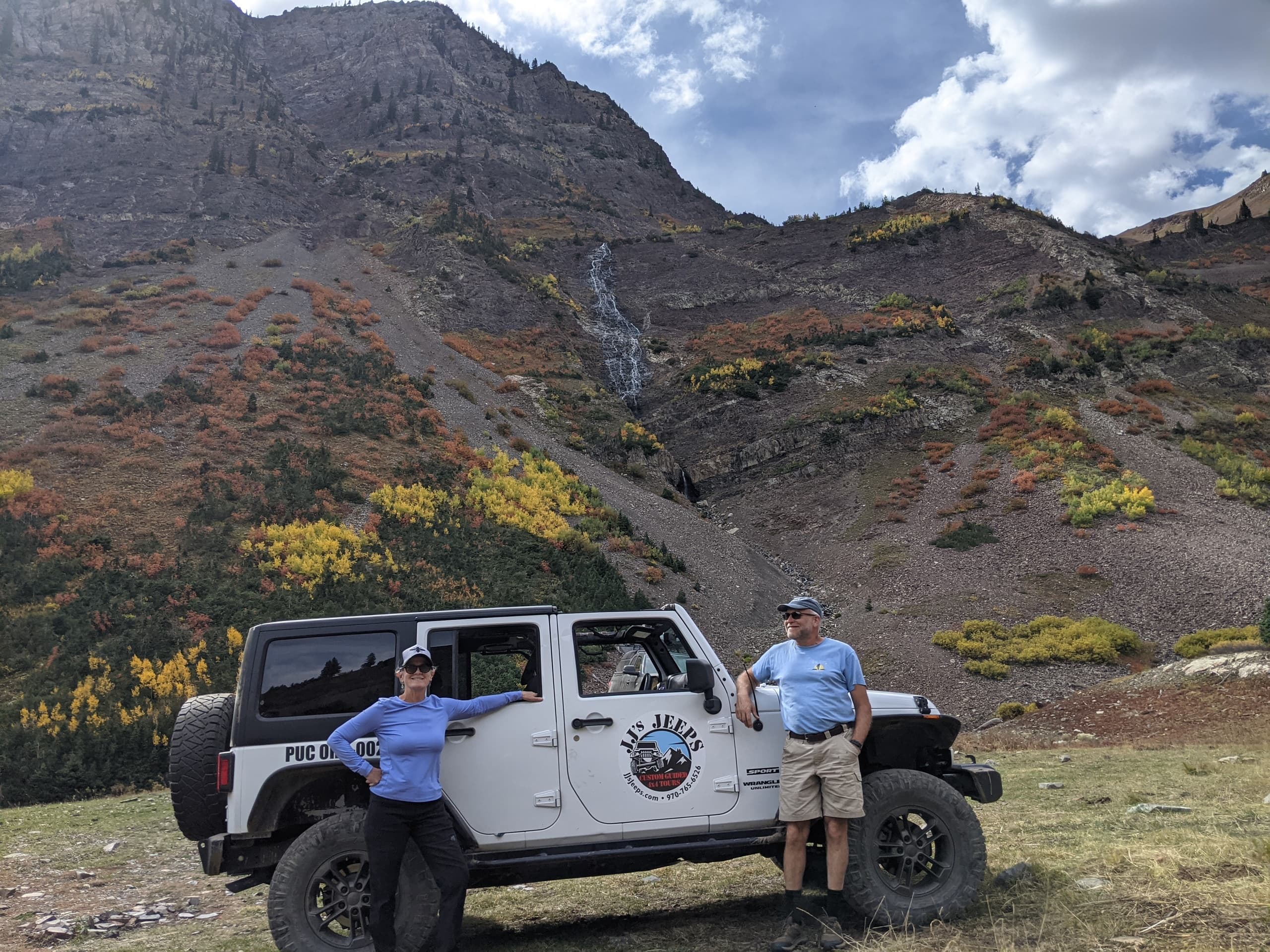 Two people posing by Jeep in high mountain valley near waterfall