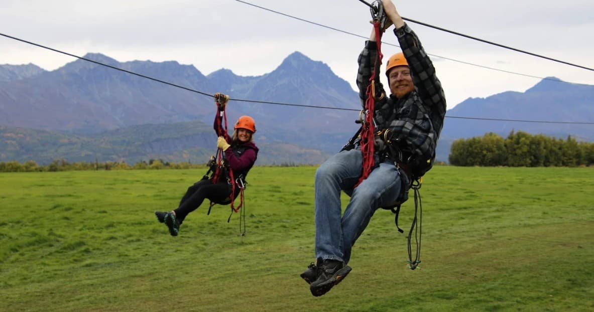 Two people ziplining over grassy field with distant mountain range